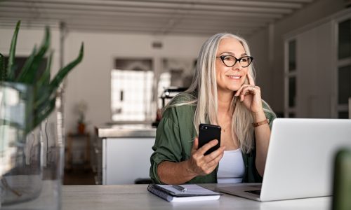 Happy senior woman holding smartphone and laptop daydreaming while looking away. Successful stylish old woman working at home while thinking about a good future. Cheerful fashionable lady entrepreneur wearing cool eyeglasses.